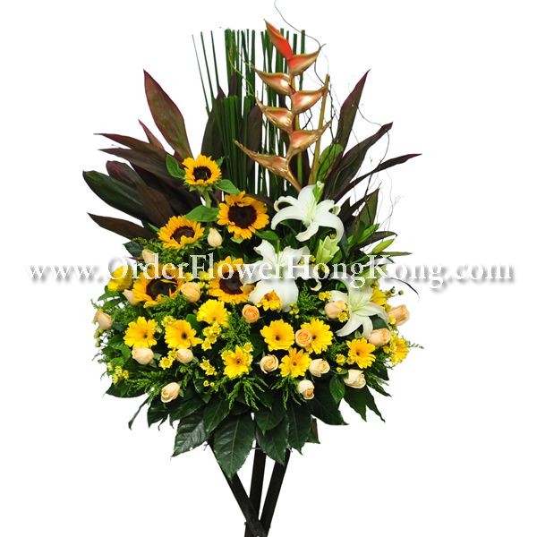 Floral arrangement with sunflowers, white lilies, roses, and tropical leaves on a stand.